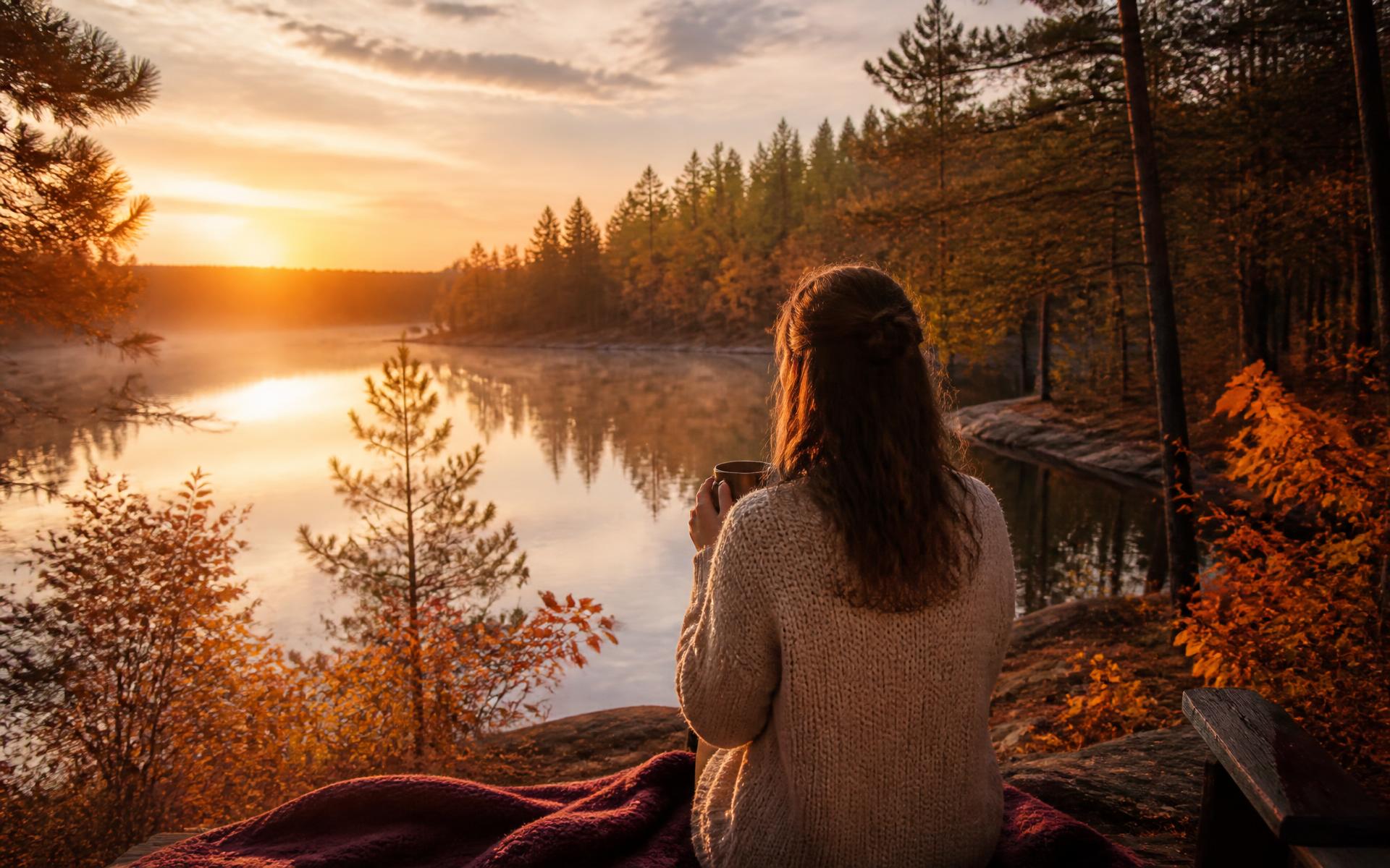 Woman sitting by a misty autumn lake at sunrise with a warm mug